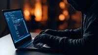 Hooded hacker typing on laptop computer with code on screen in dark room with warm bokeh lights creating cybersecurity threat atmosphere for digital security concepts.