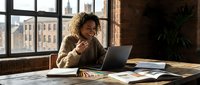 Young African American businesswoman working on laptop in modern office with city view, smiling while reviewing documents and charts on wooden desk.