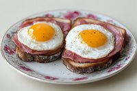 Two open-faced toasts topped with ham and sunny-side up eggs on a floral plate, served as a classic hearty breakfast meal.
