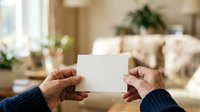 Senior adult hands holding blank white card in modern living room with natural light and home decor for greeting, invitation, or announcement messaging.