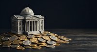 Classical government building with dome surrounded by golden and silver coins on dark wooden surface representing finance and economy concepts.