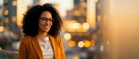 Young woman with curly dark hair and glasses wearing mustard blazer over white shirt, smiling outdoors at sunset with blurred city skyline background. Corporate professional portrait.