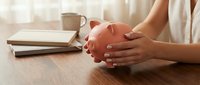 Caucasian woman hands holding pink piggy bank on wooden desk with books and mug, symbolizing savings, budgeting, and financial planning.