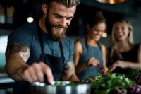 Smiling bearded chef with tattoos preparing fresh herbs in modern restaurant kitchen while customers enjoy dining experience in background.