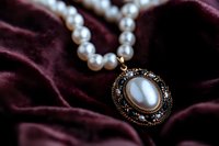 Elegant vintage pearl necklace with ornate oval pendant featuring moonstone and dark crystals, lying on luxurious burgundy velvet fabric background.