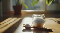 Glass jar filled with white powder or sugar on wooden table in natural sunlight with dramatic shadows and wooden spoon, minimalist food photography style.
