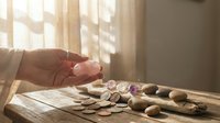 Hand holding coin above scattered coins on wooden surface with warm sunlight streaming through window creating shadows and peaceful atmosphere.
