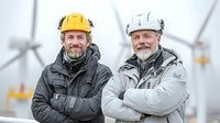 Two mature male engineers with beards wearing safety helmets standing confidently at wind farm site with turbines visible in foggy background.