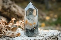 Clear quartz crystal point with natural inclusions and phantom formations standing on stone surface among autumn leaves, mystical mineral specimen in nature.