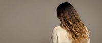 Young woman with long wavy brown hair wearing cream sweater standing with back turned against neutral beige background in minimalist studio setting.