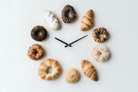 Creative clock made with assorted pastries and donuts arranged in circle on white background, representing time with bakery items.