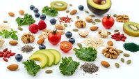 Healthy superfood ingredients scattered on white background including avocado, berries, nuts, seeds, kale and tomatoes for nutritious meal planning.