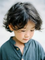 Young Asian boy with gentle smile and tousled black hair wearing blue cotton shirt, captured in soft natural light with dreamy film-like quality for lifestyle and portrait photography.