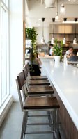 Modern cafe interior with white counter, wooden bar stools, pendant lights, and potted plants. Blurred staff and customers in background. Bright natural light from windows. Contemporary restaurant seating area.