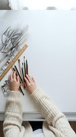 Artist hands in cream sweater arranging drawing supplies on white desk with charcoal sketch and wooden ruler for creative workspace setup.