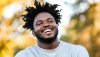 Young African American man with natural curly hair laughing genuinely outdoors, wearing light gray sweater against warm autumn bokeh background. Authentic joyful expression.