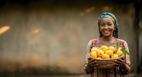 Young African female child wearing traditional blue headwrap and patterned clothing, holding wicker basket filled with fresh yellow citrus fruits, smiling at camera against blurred rustic background.