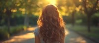 Young woman with long wavy brown hair viewed from behind at sunset in park. Golden sunlight creates dreamy backlit effect through flowing locks against blurred nature background.