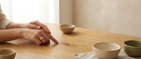 Woman hands resting on wooden table with ceramic bowls in minimalist kitchen interior. Natural lighting creates calm atmosphere for mindful living.