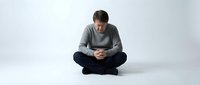 Middle-aged Caucasian man sitting cross-legged on floor, head bowed, hands clasped, expressing sadness, loneliness, or deep thought.
