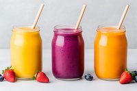 Colorful fresh smoothies in mason jars with straws. Yellow mango, purple berry and orange fruit drinks garnished with strawberries and blueberries on white surface.