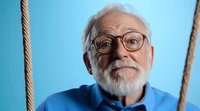 Cheerful elderly Caucasian man with white beard and glasses smiling warmly in blue shirt against turquoise background for lifestyle and aging concepts.