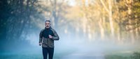 Middle-aged Caucasian man jogging through misty forest path during golden hour sunrise, wearing black athletic wear for healthy lifestyle and fitness.