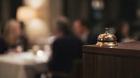 Vintage brass service bell on wooden counter in upscale restaurant with blurred diners in background creating warm hospitality atmosphere.