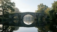 Ancient stone bridge with perfect reflection in calm water surrounded by autumn trees creating symmetrical mirror image in peaceful countryside setting.