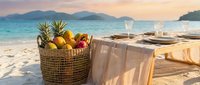 Tropical beach picnic setup with fresh pineapples and fruits in wicker basket on sandy shore during golden sunset with turquoise ocean and mountains.
