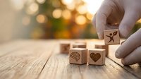 Hand placing wooden cube with leaf symbol next to heart and brain icons on rustic table with warm bokeh background for eco-friendly business concepts.