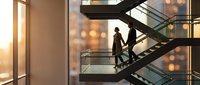 Professional businesswoman in elegant suit ascending modern glass staircase in contemporary office building during golden hour with city lights backdrop.