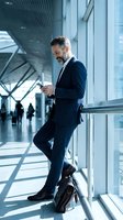 Confident businessman using smartphone in modern office building corridor with natural light and glass windows for corporate communication.