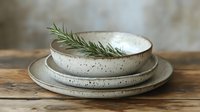 Handmade ceramic dinnerware set with natural speckled glaze and fresh rosemary sprig on rustic wooden table for organic kitchen styling and tableware.