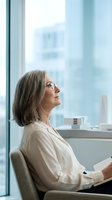 Mature woman with gray hair and glasses sitting by office window, holding tablet, gazing at cityscape view for business, corporate, and professional concepts.