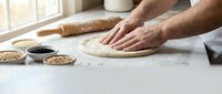 Male baker hands kneading fresh dough on floured kitchen counter with rolling pin and ingredients for homemade bread making process in warm natural light.