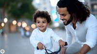 Happy father teaching young son to ride bicycle on city street during evening with bokeh lights in background.