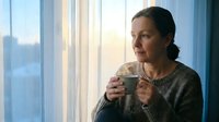 Middle-aged Caucasian woman in cozy sweater drinking coffee by window, contemplating morning light in peaceful home setting for lifestyle content.