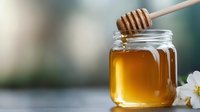 Golden honey dripping from wooden dipper into glass jar with white flowers on blurred background for natural sweetener and organic food concepts.
