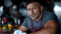Middle-aged Hispanic male chef with apron looking thoughtfully at camera, fresh produce on counter in restaurant kitchen background.