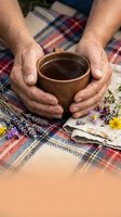 Elderly hands holding wooden bowl with herbal tea on plaid blanket with wildflowers and dried herbs for natural wellness and traditional medicine.