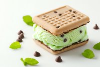 Mint chocolate chip ice cream sandwich with chocolate cookie wafers, garnished with fresh mint leaves and chocolate chips on white background.