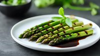 Fresh green asparagus spears arranged on white ceramic plate with balsamic reduction, garnished with micro herbs against dark background, food styling.