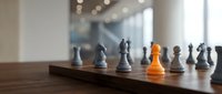 Chess pieces on wooden board with orange king standing out from black pieces in modern office setting with blurred background lights.