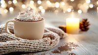 Hot chocolate topped with whipped cream and cocoa powder in white mug, cozy winter setting with knit scarf, glowing candle and pine cone against bokeh lights.