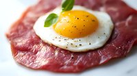 Sunny side up egg with bright yellow yolk served on thin sliced prosciutto ham garnished with fresh basil leaf and black pepper on white plate, macro food photography.