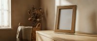 Empty wooden picture frame on dresser in warm sunlit bedroom interior with dried flowers and soft natural lighting for home decor and design mockup.