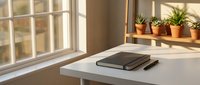Minimalist home office workspace with laptop on white desk near window with natural sunlight and small potted plants on wooden shelf creating productive atmosphere.