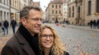 Happy mature couple embracing on cobblestone street in historic European city center during autumn day with warm sunlight and blurred pedestrians in background.