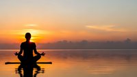 Silhouette of person meditating in lotus pose on paddleboard at sunrise over calm water with golden sky reflection for wellness and mindfulness concepts.